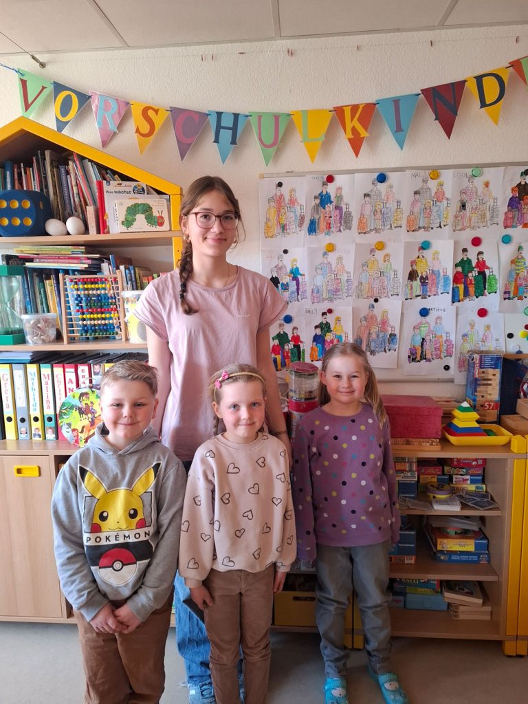 Eine junge Erzieherin mit Brille und einem rosafarbenen Shirt steht vor einer Wand mit Bücher- und Spieleregal, Zeichnungen und einer Wimpelkette, auf der das Wort "Vorschulkinder" zu lesen ist. Vor ihr stehen zwei Mädchen und ein Junge. Alle vier Personen auf dem Bild lächeln und freuen sich.