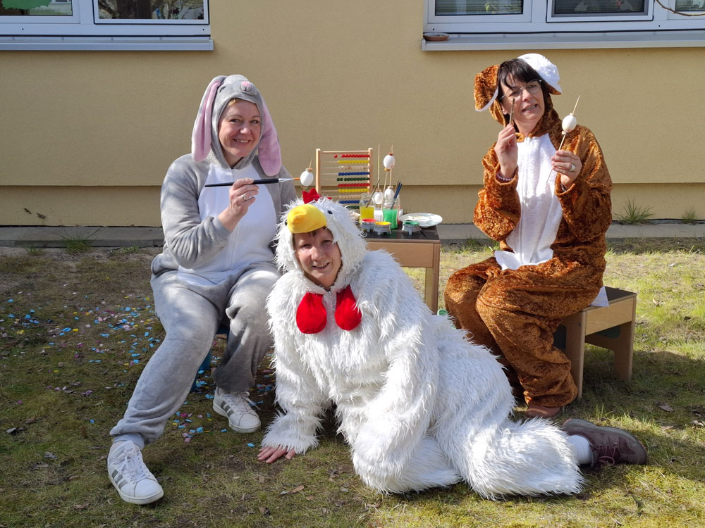 Zu sehen sind drei Erzieherinnen, die an einem kleinen Tisch sitzen und Ostereier für die Kamera bemalen. Ein Rechenschieber steht ebenfalls auf dem kleinen Tisch. Eine der Erzieherinnen trägt ein Ganzkörper-Hühnerkostüm und posiert vor dem Tisch auf dem Rasen. Die beiden anderen Erzieherinnen, halten ein jeweils einen Pinsel und ein unbemaltes Ei in der Hand. Beide tragen ein Ganzkörper-Hasenkostüm. Eins ist hellgrau, eins ist braun.